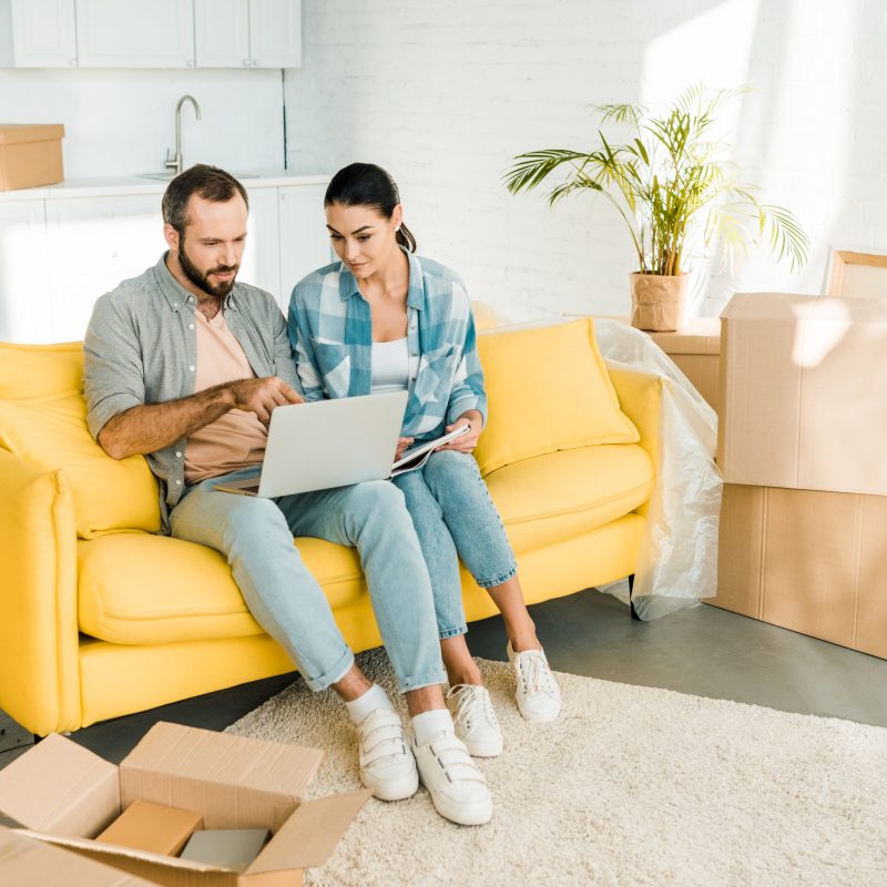focused couple sitting on couch, using laptop and planning relocation to new house, moving concept