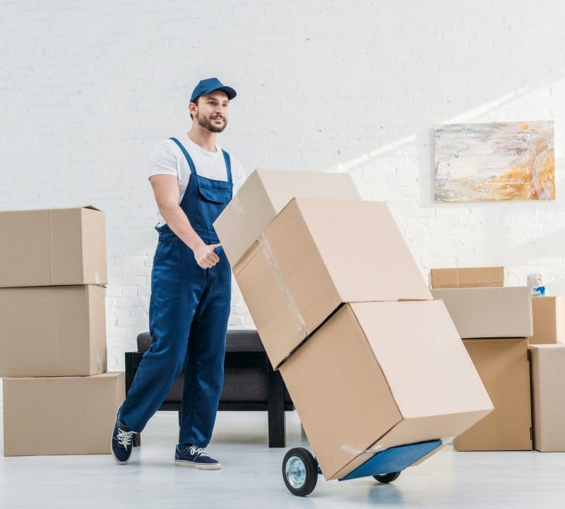 mover-in-uniform-transporting-cardboard-boxes-on-hand-truck-in-apartment.jpg