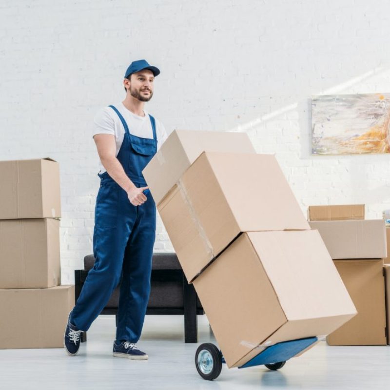 mover-in-uniform-transporting-cardboard-boxes-on-hand-truck-in-apartment.jpg